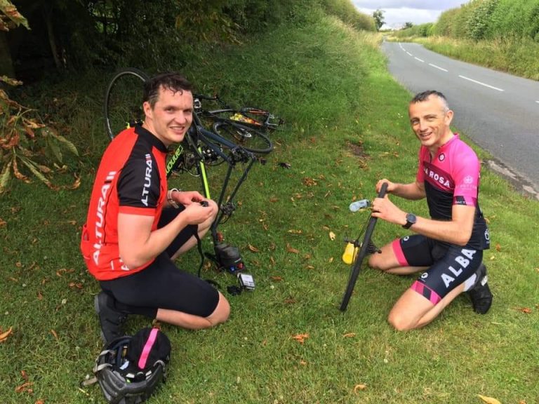 Lending a hand, and giving some knowledge... Two cyclists kneeling on a grassy bank, one helping the other repair a puncture.