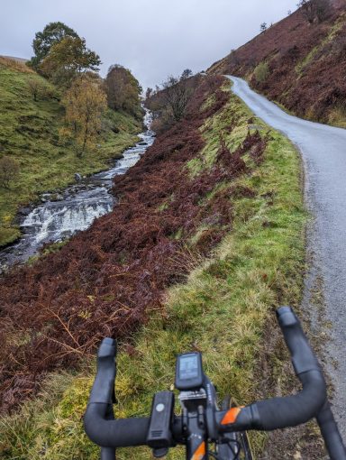 Wales View from a bike pointing up a hill with a stream on one side and heather covered roadside on the other.