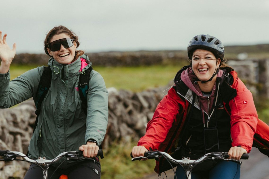 Two ladies riding their bikes on a road in front of a dry stone wall. One is waving, both are smiling, and wearing comfortable clothes.