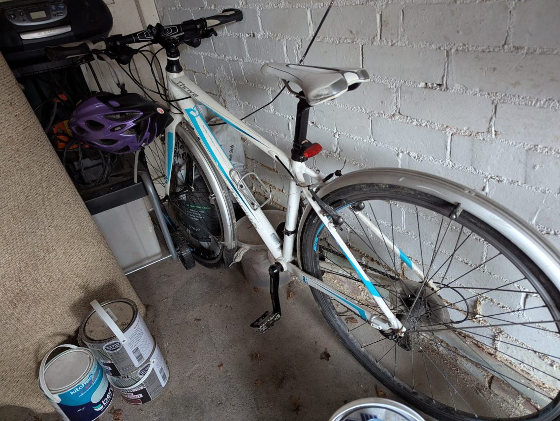 A white bike, sat in the corner a garage surrounded by paint cans, an old carpet and it's obviously rarely used.