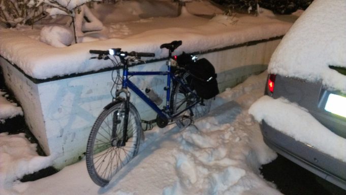 Old Lump An old hybrid bike, with panniers on, leaning against a wall on a very snowy evening. The lights are still lit from being ridden home.