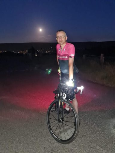After Everesting A tired cyclist smiling while stood over a bike. The moon is out in the background; it was taken at 10pm in the middle of summer.